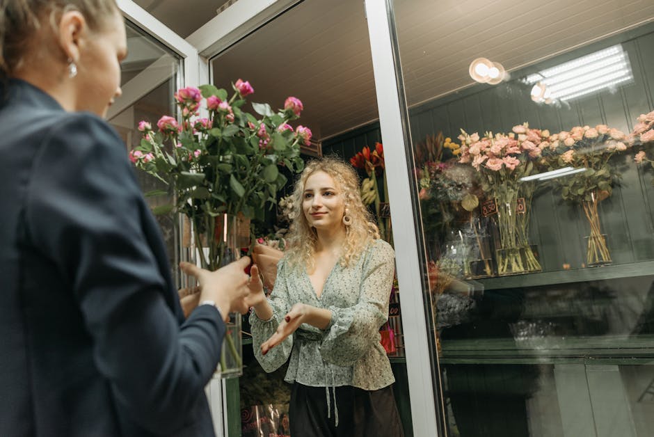 A female florist inside a flower shop on Cambridge Road is handing a bouquet of pink roses with green foliage to a customer. The bouquet is wrapped in brown craft paper and placed in a tall glass vase