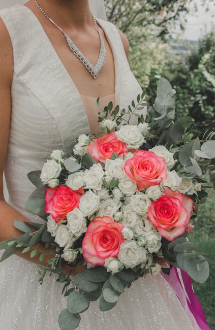 A bride holding a large floral bouquet featuring vibrant pink roses surrounded by smaller white roses and silver-green eucalyptus leaves. She is wearing a white wedding gown with subtle glitter accent
