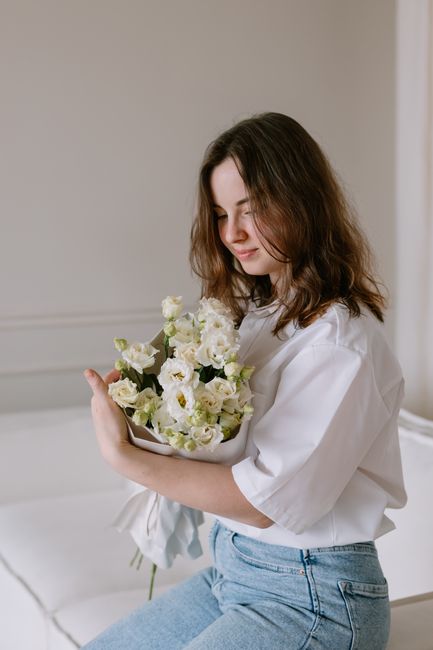 A young woman with shoulder-length brown hair, wearing a white blouse and light blue jeans, is gently holding a bouquet of fresh white flowers including lisianthus and roses, with green foliage, wrapp