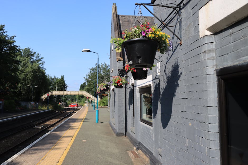 A flower basket filled with a vibrant assortment of pink, yellow, purple, and white flowers, including petunias and geraniums, hanging from a black metal bracket attached to the grey brick exterior wa