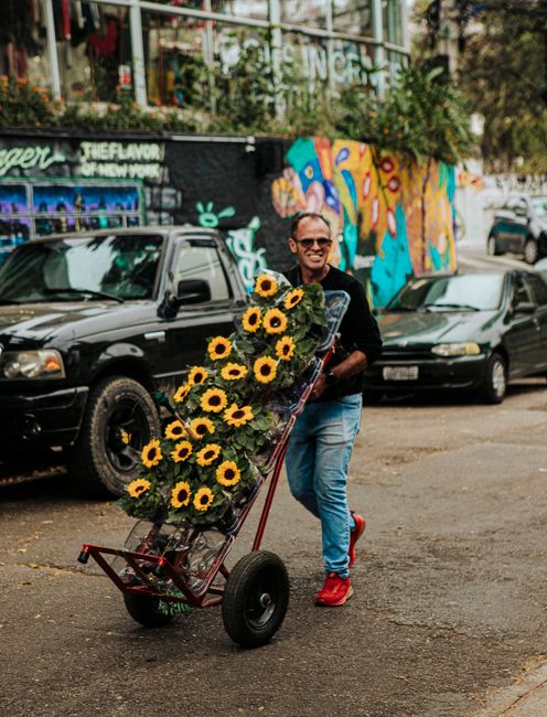 A man standing outdoors, holding a large floral arrangement in a red hand truck filled with vibrant yellow sunflowers and green foliage. He is smiling and wearing sunglasses, a dark shirt, blue jeans,