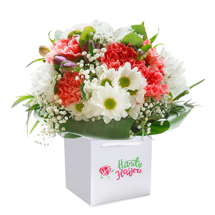 Tall and immaculate, this floral arrangement stands in a striking white square gift bag-its artistry telling a story of Norbiton's signature style and community pride. The bouquet bursts forth with plush, ruffled carnations in coral, peach, and vibrant red, each bloom perfectly unfurled to catch the light and fill the air with gentle fragrance. Bright white daisies punctuate the composition, their lively lime-green centers reminiscent of sunlit meadows near Norbiton's Canbury Gardens, bringing a lively freshness. Wisps of white baby's breath, their tiny blossoms echoing soft clouds, intermingle throughout, creating a dreamlike softness that dances between the larger blooms. Punctuating this tapestry, a few tightly curled, purple-tinged buds peek from the left edge, hinting at the bouquet's evolving beauty. Enveloping the arrangement, deep green, lustrous foliage adds drama and richness, its elegant leaves forming a natural cradle for the display. The understated white gift bag, complete with white ribbon handles, underscores the bouquet's refined presentation and practical appeal for local deliveries-ideal for gifting on any milestone, from anniversaries to congratulations. Set against a pure white background, every floral detail appears crisp, vivid, and thoughtfully arranged, embodying the care and expertise Norbiton florists bring to every creation. This bouquet captures the essence of giving-a moment of beauty, freshness, and possibility hand-delivered right to a neighbour's door.