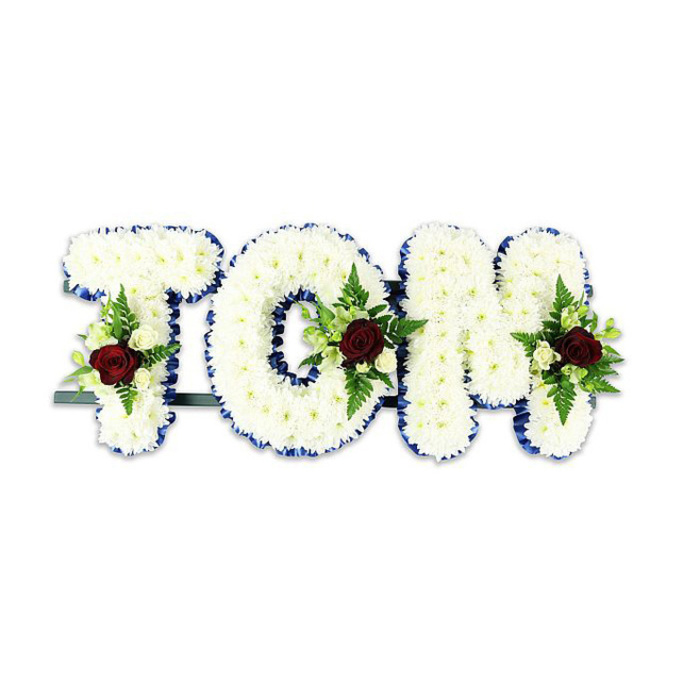 A striking bespoke sympathy piece spells TOM in bold, three-dimensional letters, each letter densely covered with innumerable small white chrysanthemum blooms to create a uniform, soft-textured surface. The edges of every letter are crisply finished with a royal blue ribbon which provides a vivid, contrasting frame to the pale blooms and gives each character a graphic clarity against the stark white background. Placed thoughtfully on the centre or lower portion of each letter is a modest cluster of accent flowers: a single deep red rose serves as the focal point for each spray, supported by smaller creamy white spray roses and lengths of green fern fronds and leafy foliage that add sculptural depth and natural movement. A thin dark green or black metal bar is visible beneath the arrangement, indicating the support structure that allows the letters to be displayed upright at a funeral venue or graveside. The composition balances formality and tenderness - the chrysanthemums suggest purity and resilience while the red roses quietly signify love and remembrance - and emits a restrained floral scent, the rose lending a soft warmth to the otherwise cool palette. Crafted with attention to proportion and finish for memorial services in Norbiton and neighbouring districts such as Kingston Hill and Coombe, this name tribute is designed to read clearly from a distance while offering thoughtful detail up close for those who come to pay their respects.