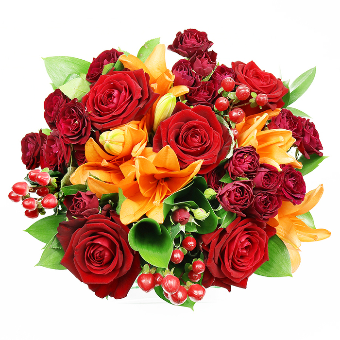Captured from above on a stark white surface, this lavish, circular bouquet is a study in warm contrasts and floral texture. A handful of large, fully opened deep red roses command attention at the centre, their velvety petals arranged in dense spirals that look both sumptuous and inviting. Radiating inward and outward from those anchors are bright orange Asiatic lilies-some with wide, trumpet-like faces and visible stamens, others still coyly unfurling-adding structural drama and a citrus-spiced lift to the scent profile. Intermingled close to the roses are compact spray roses in a wine or burgundy tone; their miniature blooms create a plush, layered depth that balances the boldness of the larger flowers. Glossy hypericum berries appear in small clusters across the surface, providing playful, rounded punctuation that reflects light and invites touch. Lush green foliage-broad, glossy leaves similar to ruscus or salal-emerges between the blooms to give the design breathing room and a fresh contrast against the reds and oranges. The bouquet's lighting is gentle and even, highlighting the satiny sheen of petals and the berry gloss without casting distracting shadows. This arrangement feels celebratory and intimate at once, suited to anniversary gestures, autumnal décor, or a heartfelt thank-you. I imagine it delivered through Norbiton's neighbourhood streets, perhaps placed on a windowsill overlooking local high-street bustle or received after a walk past Kingston Market-an elegant blend of warmth, texture, and floral craftsmanship.