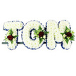 This photograph shows a personalised floral name tribute reading TOM, created as three-dimensional letters for a funeral or memorial service close to Norbiton. Each capital letter is completely covered in pristine white chrysanthemums, their tightly packed, ruffled petals forming a thick, cushiony layer that softens the strong, blocky shapes of the word. Around the edge of each letter, a royal blue satin ribbon is carefully pinned, the smooth, slightly glossy fabric catching subtle highlights and providing a clear, defined outline that frames the white blooms. At specific points-the upper left of the T, the curve of the O and the top right of the M-small, elegant floral sprays interrupt the white surface. These are composed of deep red roses with velvety petals, smaller cream or white spray roses and slim, white freesia-type flowers, all cradled in fresh green foliage and delicate fern leaves that spill gently over the chrysanthemums. The letters rest in a row on a slim, dark metal frame, positioned so they could easily be displayed on a stand at a Norbiton church or in a chapel of rest. The combination of pure white, rich red, cool blue and lush green gives the piece a calm, reflective presence, expressing purity, love and remembrance. It is the sort of carefully considered tribute chosen by families around Norbiton who want their flowers to convey respect and affection for Tom when words alone are not enough.
