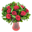 A lush, rounded bouquet of a dozen or more deep crimson roses fills this image, each bloom opened to reveal velvety, spiralling petals that graduate from a wine-dark core to a glowing crimson edge. The heads are tightly clustered into a classical dome, their plush texture catching soft, even studio light so the fine ridges and velvet sheen of each petal are visible. Nestled among the roses is an abundant palette of dark green foliage: flat, glossy leaves-suggestive of Ruscus or Salal-form a dense, verdant backdrop, while larger emerald leaves have been deliberately rolled and curled into sculptural spirals that arc around and cradle the blooms. Those artful, almost architectural leaf loops lend a modern twist to an otherwise timeless composition and create depth and negative space that makes each rose read clearly against the white field.
The bouquet sits in a tall, slender translucent ruby-red glass vase whose subtle twisted or ribbed surface throws back vertical highlights and mirrors the intensity of the petals, anchoring the arrangement in colour and line. The vase's narrow waist and stable base are hinted at beneath the foliage, giving a sense of elegant balance and height. Against the crisp white background every tone-from the deepest crimson to the glossy forest greens-pops with dramatic clarity, and the overall effect reads as both romantic and refined.
As a local florist's image leading into ideas for anniversaries or Valentine's Day, the picture evokes warm fragrance, classic devotion and considered craftsmanship-an arrangement you might imagine delivered to a Norbiton home near Norbiton station or the Kingston riverside to mark a quiet, intimate celebration.
