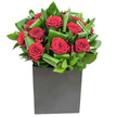 A compact, modern arrangement that speaks with quiet assurance: deep, velvety red roses clustered at the centre like small, passionate embers, each bloom opening in layered spirals of plush petals. Around them, broad glossy green leaves have been folded and curled with deliberate care, their waxy sheen catching the light and tracing a rhythmic pattern of loops and arcs that gives the composition a sense of movement and architectural balance. The flowers sit firmly in a matte black square vessel, its clean geometric lines providing a bold, grounded base that offsets the organic softness above. The studio lighting is clear and even, making the crimson tones glow with a velvet depth while throwing crisp highlights across the leaves so textures read almost tactilely. If you lean in, there is the imagined scent of freshly cut rose, pure and classic, warmed by the suggestion of green, leafy freshness rather than sweetness alone. As a florist I picture this piece set on a contemporary office desk in central Norbiton or as a table centre for an intimate anniversary supper near Canbury Gardens - it adds both drama and restraint. The design favours clean silhouettes and refined contrast rather than fussy embellishment, and its confident simplicity makes it suitable for declarations of love, meaningful congratulations, or as a striking focal point to lift a living room. Every fold of foliage and each measured bloom conveys considered craftsmanship, a studied restraint that nevertheless rewards close attention with a quietly luxurious presence.